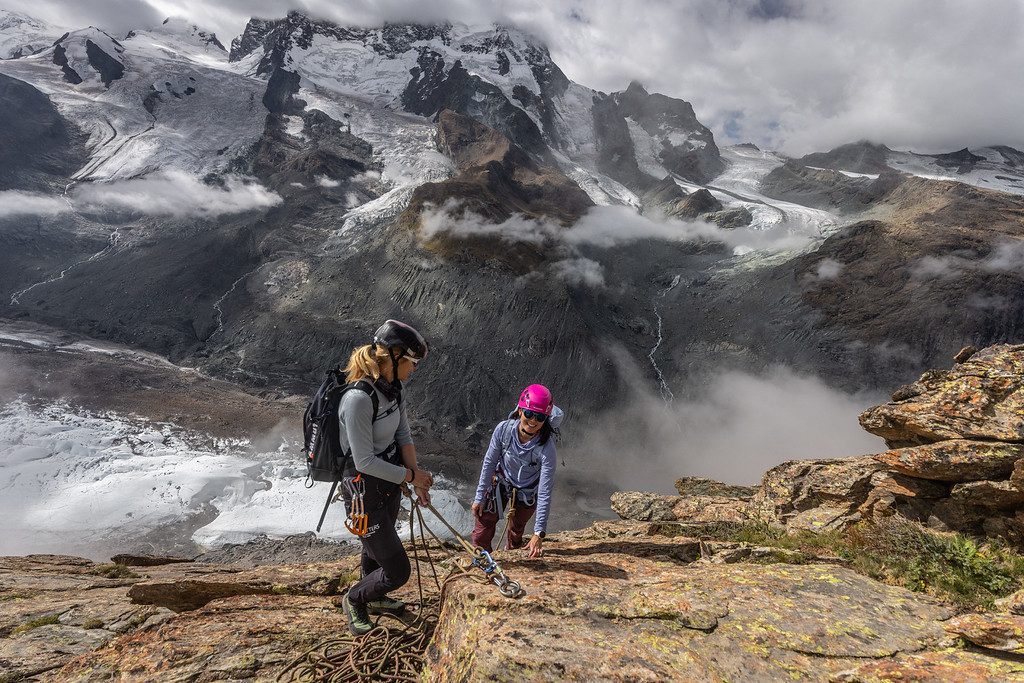 Two women in helmets and harnesses climbing a rocky ridge above a glacier on the Matterhorn.