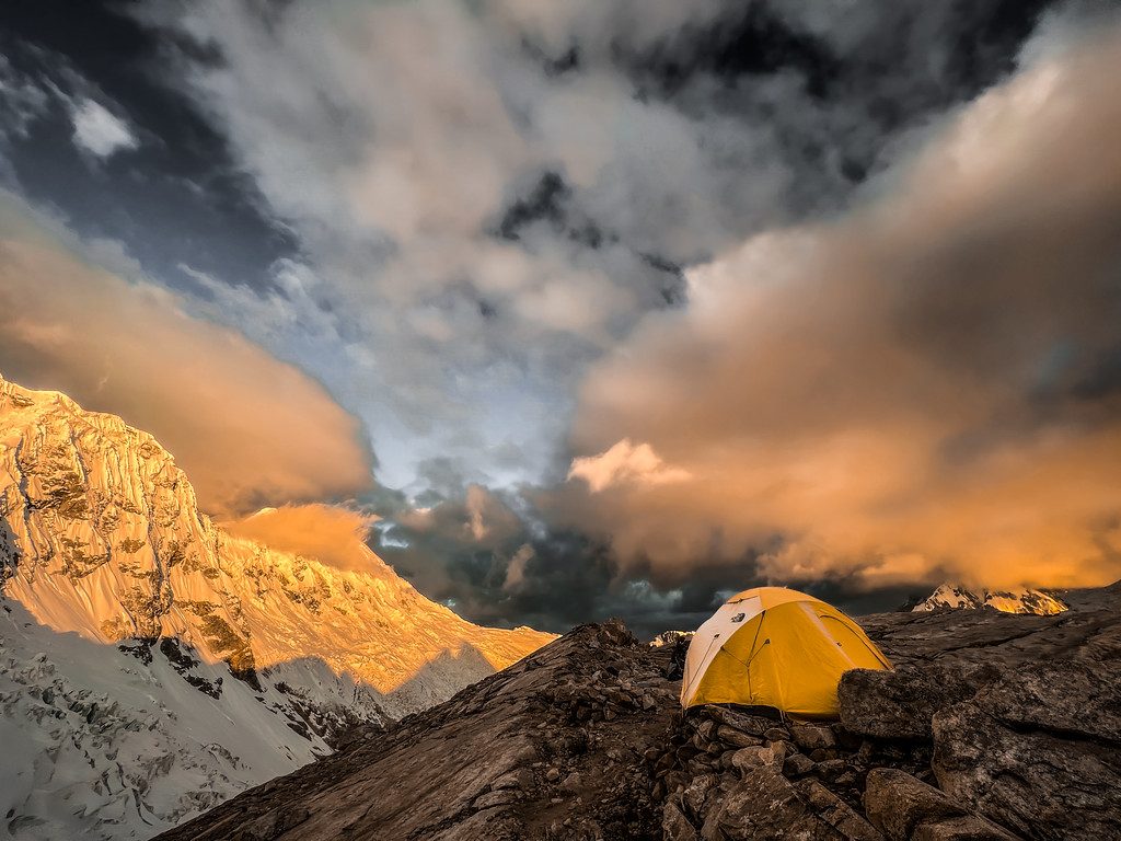 A yellow tent on a rocky ridge at sunset with a glowing peak and storm clouds behind it.